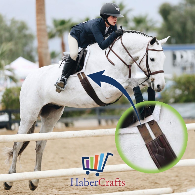 May include: A white horse jumping over a white fence with a rider wearing a black jacket and a helmet. The horse is wearing a brown leather saddle and a brown leather girth with a white stripe.