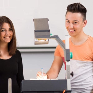 May include: A young woman with long brown hair is standing next to a young man with short brown hair. They are both looking at the camera. The man is holding a pen and is pointing at a projector. The projector is on a table and is pointed at a white wall.