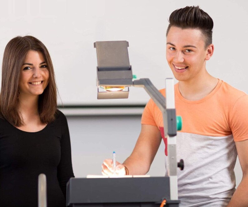 May include: A young woman with long brown hair is standing next to a young man with short brown hair. They are both looking at the camera. The man is holding a pen and is pointing at a projector. The projector is on a table and is pointed at a white wall.