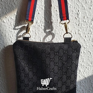 May include: Black crossbody bag with a black and white GG pattern. The bag has a red and blue adjustable strap with gold hardware.