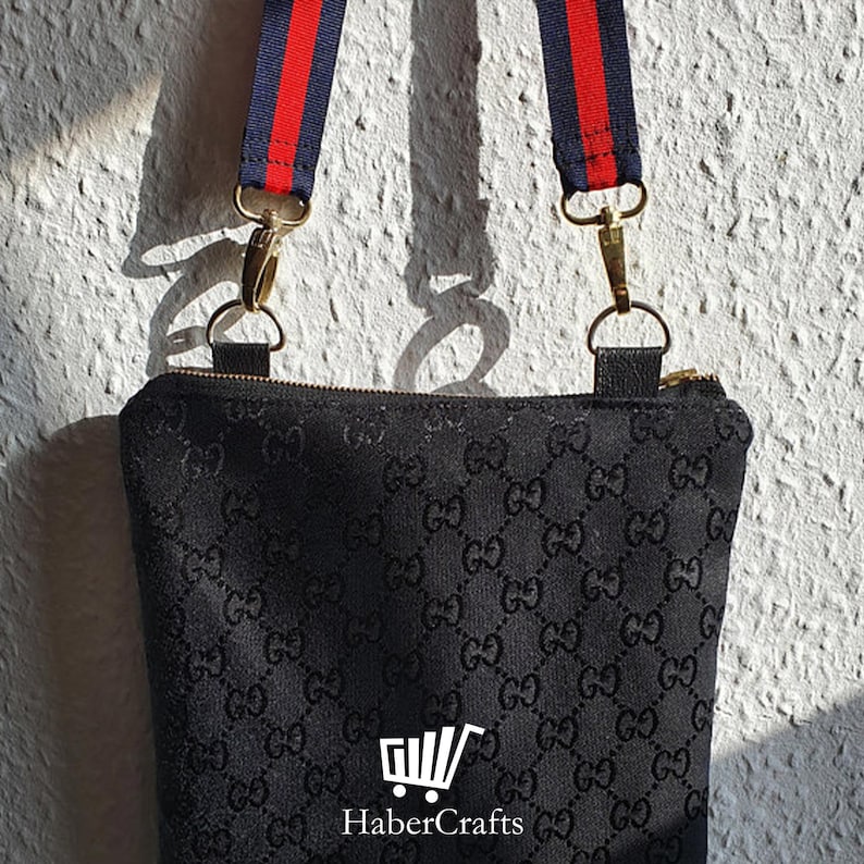 May include: Black crossbody bag with a black and white GG pattern. The bag has a red and blue adjustable strap with gold hardware.