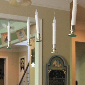 May include: Five white battery-operated candles hanging from the ceiling with gold candle holders. The candles are suspended from the ceiling with clear fishing line. The candles are in a room with a light brown ceiling and white walls. There is a sign on the wall that says "Do solemnly swear that I am up to no good."