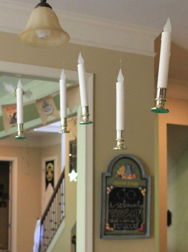 May include: Five white battery-operated candles hanging from the ceiling with gold candle holders. The candles are suspended from the ceiling with clear fishing line. The candles are in a room with a light brown ceiling and white walls. There is a sign on the wall that says "Do solemnly swear that I am up to no good."