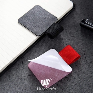 May include: A notebook with a black leather cover and a black elastic pen holder. A purple adhesive note with a white peel-off section and a red elastic band is attached. The HaberCrafts logo is visible.