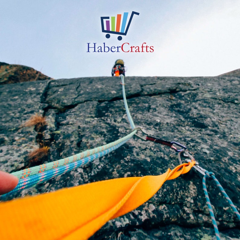 May include: A close-up of a rock climbing rope with a yellow climbing sling and a carabiner. The rope is blue, green, and pink. The rope is attached to a rock face.