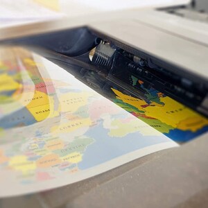 May include: A close-up of a flatbed scanner with a colorful world map being scanned. The map is yellow, blue, green, and red. The scanner is white and gray.