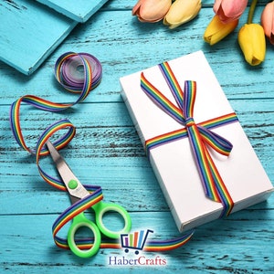 May include: A white gift box wrapped with rainbow ribbon, a pair of green scissors, and rainbow ribbon on a blue wooden surface. The ribbon is tied in a bow on top of the box. There are also some colourful flowers in the background.