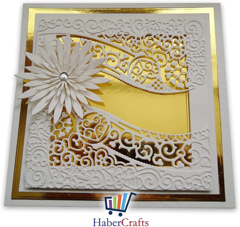 K&ouml;nnte beinhalten: Eine wei&szlig;e Karte mit goldfarbenem Rand und einem wei&szlig;en Papier-Serviettenmuster. Das Serviettenmuster hat eine wei&szlig;e Blume mit einem Strassstein in der Mitte oben links. Die Karte ist leer und bereit f&uuml;r eine Nachricht.
