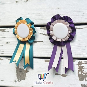 May include: Two blank rosette ribbons with a white centre circle. The rosette on the left has teal and gold ribbons. The rosette on the right has purple and beige ribbons.
