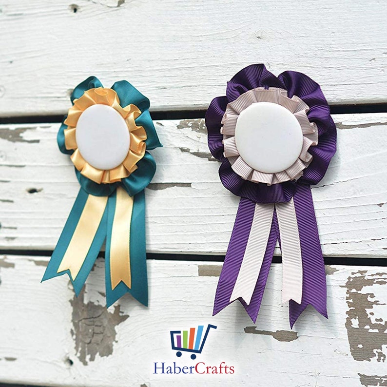 May include: Two blank rosette ribbons with a white centre circle. The rosette on the left has teal and gold ribbons. The rosette on the right has purple and beige ribbons.