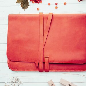 May include: A bright red leather laptop sleeve with a strap closure. The sleeve is on a white wooden surface.
