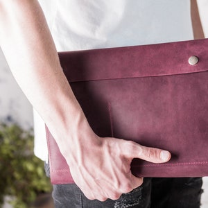 May include: A burgundy leather laptop sleeve with a snap closure. The sleeve has a simple design with a stitched edge and a small pocket on the front. The leather has a slightly distressed look.