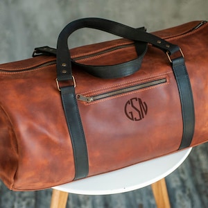 Może przedstawiać: Brązowa sk&oacute;rzana torba podr&oacute;żna z czarnymi sk&oacute;rzanymi paskami i monogramem "GSN" z przodu.