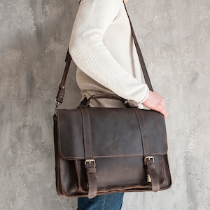 May include: A brown leather briefcase with a strap and buckles. The briefcase is being held by a person.
