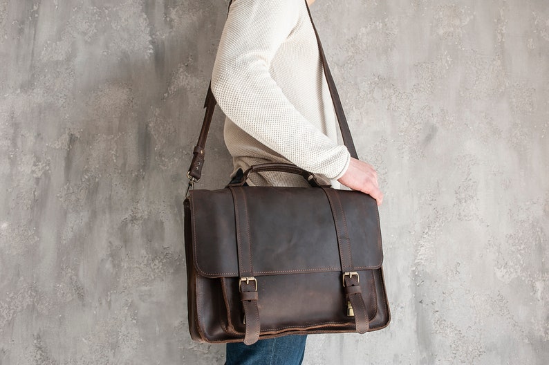 May include: A brown leather briefcase with a strap and buckles. The briefcase is being held by a person.