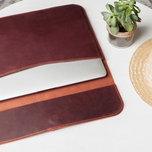 May include: A brown leather laptop sleeve with a flap that folds over to close. The sleeve is open and a laptop is inside. The sleeve is on a white surface with a small potted plant and a straw hat.