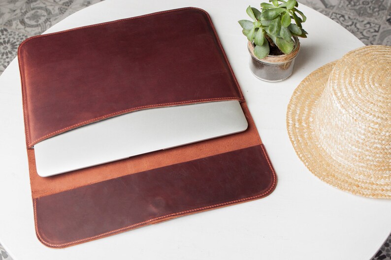May include: A brown leather laptop sleeve with a flap that folds over to close. The sleeve is open and a laptop is inside. The sleeve is on a white surface with a small potted plant and a straw hat.