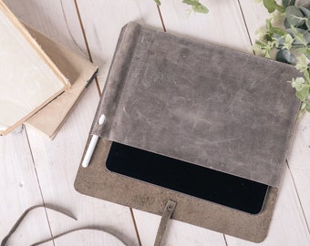 May include: A gray leather tablet sleeve with a white stylus and a black tablet inside. The sleeve has a strap and a flap that closes with a button.