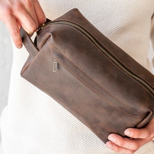 May include: A brown leather toiletry bag with a zipper closure and a small handle. The bag is rectangular and has a textured appearance. The zipper is a contrasting color, and there is a small metal tag on the side.