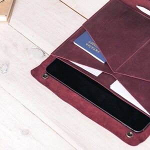May include: Burgundy leather folio with multiple pockets, holding a tablet, documents, and a passport. A pen and stylus are secured in a loop. The folio is open, revealing its contents on a light wood surface.
