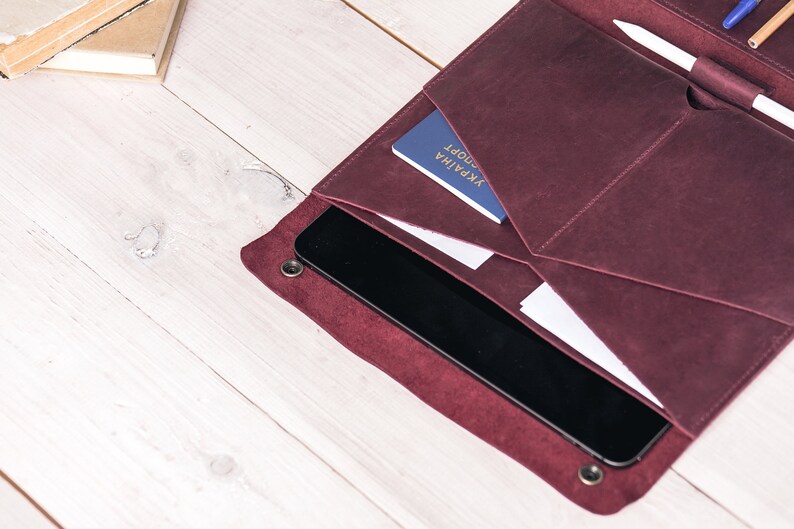 May include: Burgundy leather folio with multiple pockets, holding a tablet, documents, and a passport. A pen and stylus are secured in a loop. The folio is open, revealing its contents on a light wood surface.