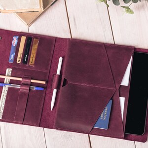 May include: Burgundy leather folio with multiple pockets for cards, cash, pens, and a tablet. The folio is open, revealing a tablet, credit cards, a pen, a pencil, and a passport. The folio is on a white wooden surface.