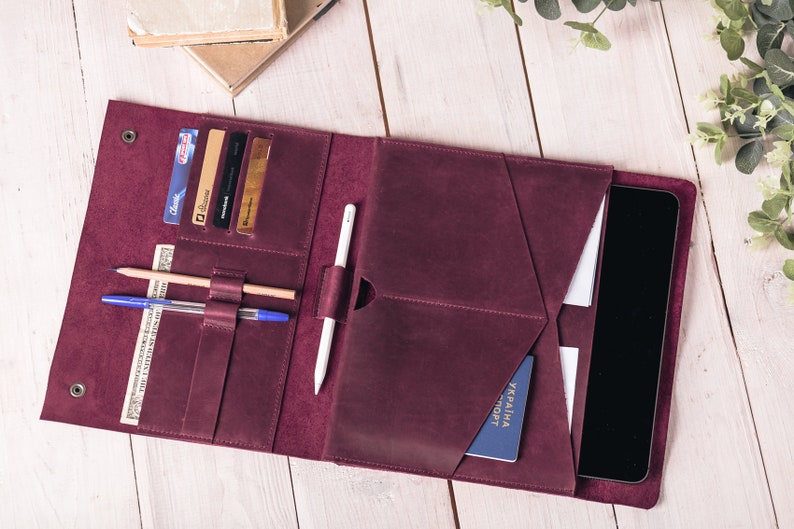May include: Burgundy leather folio with multiple pockets for cards, cash, pens, and a tablet. The folio is open, revealing a tablet, credit cards, a pen, a pencil, and a passport. The folio is on a white wooden surface.