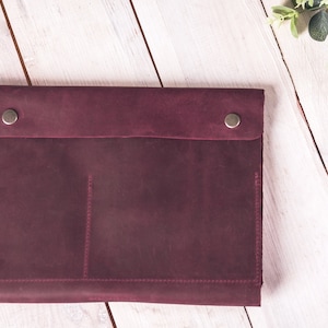 May include: A burgundy leather laptop sleeve with a flap closure secured by two silver-toned snap buttons. The sleeve has a pocket detail and is stitched with burgundy thread. The background is a white wooden surface.