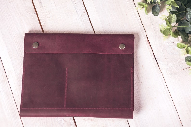 May include: A burgundy leather laptop sleeve with a flap closure secured by two silver-toned snap buttons. The sleeve has a pocket detail and is stitched with burgundy thread. The background is a white wooden surface.