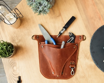 Può includere: Una cintura portautensili in pelle marrone con più tasche e un fermaglio in metallo. La cintura è piena di utensili, tra cui una lima, una pinza, un cacciavite e una chiave.