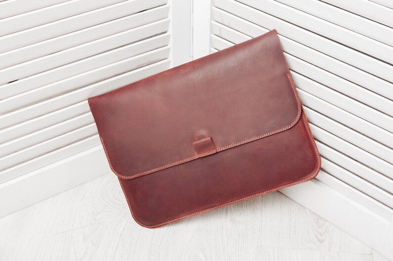 May include: A brown leather laptop sleeve with a flap closure and a stitched edge.