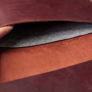 May include: A brown leather laptop sleeve with a gray felt lining. The sleeve is open and the felt lining is visible.
