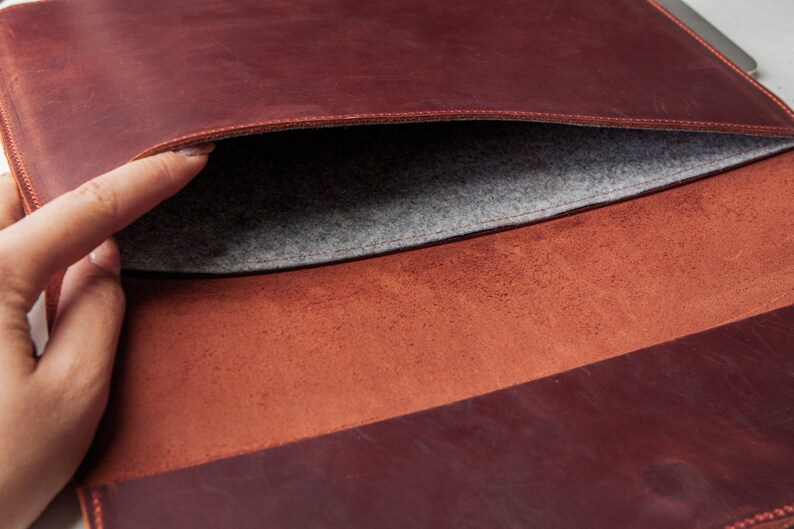 May include: A brown leather laptop sleeve with a gray felt lining. The sleeve is open and the felt lining is visible.