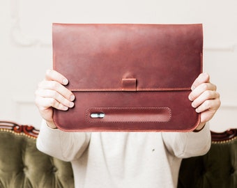 May include: A brown leather laptop sleeve with a flap closure and a pen holder. The sleeve is held in front of a person's face.