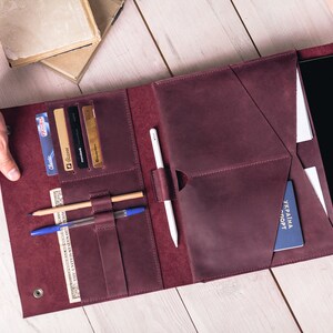 May include: A burgundy leather folio with multiple pockets and compartments. It holds a tablet, cards, pens, and a passport. The folio is open, revealing its contents, and is held by two hands.