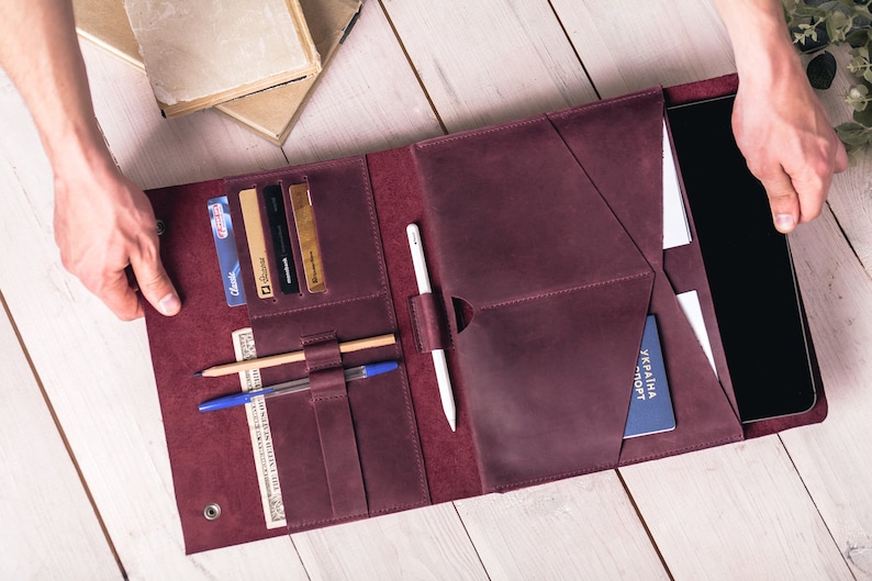 May include: A burgundy leather folio with multiple pockets and compartments. It holds a tablet, cards, pens, and a passport. The folio is open, revealing its contents, and is held by two hands.
