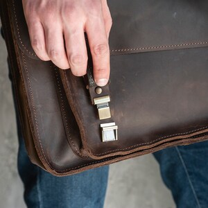 May include: A brown leather briefcase with a metal clasp and a strap. The briefcase is closed and has a flap that is folded over.