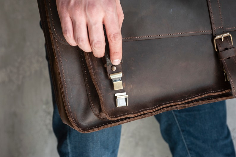 May include: A brown leather briefcase with a metal clasp and a strap. The briefcase is closed and has a flap that is folded over.