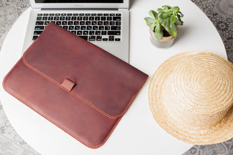 May include: A brown leather laptop sleeve with a flap closure and a small strap. The sleeve is on a white table with a laptop, a potted plant, and a straw hat.