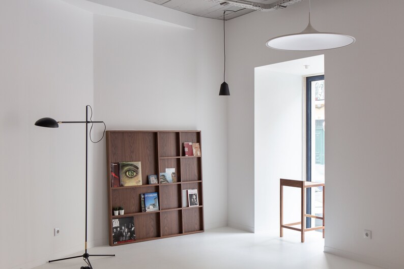 May include: A white room with a wooden bookshelf filled with books and magazines. There is a black floor lamp in the corner of the room and a white ceiling light fixture hanging from the ceiling. A small wooden stool is in front of a window.