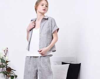 May include: A woman wearing a gray linen shirt with a white tank top underneath and matching gray linen pants. She is standing in a white room with a white chair and pillows.