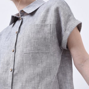 May include: A gray linen shirt with a button-up front and a single chest pocket. The shirt has short sleeves with rolled cuffs.
