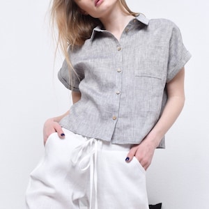 May include: A gray linen shirt with a button-up front and short sleeves. The shirt has a pocket on the left side and is worn with white linen pants.