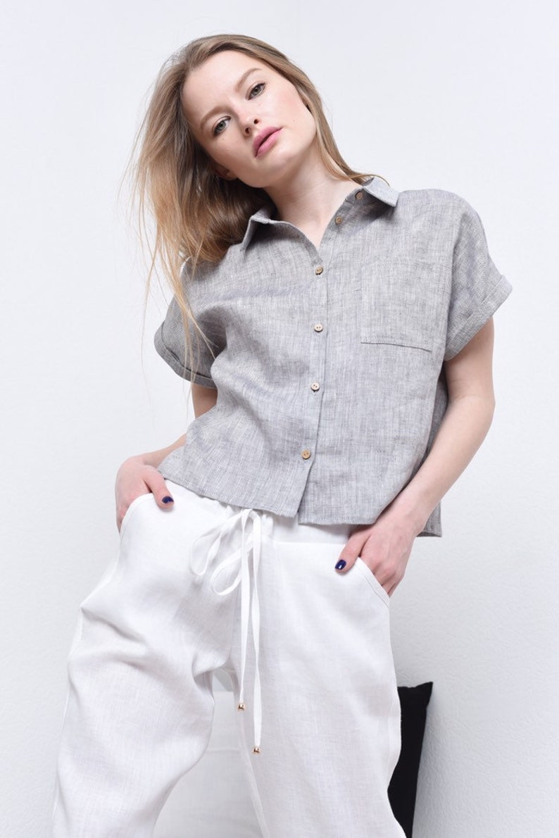 May include: A gray linen shirt with a button-up front and short sleeves. The shirt has a pocket on the left side and is worn with white linen pants.