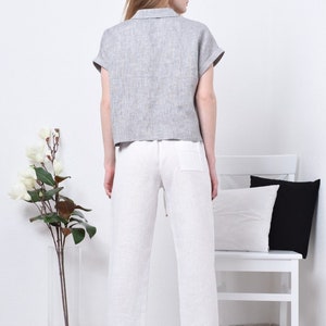 May include: A woman wearing a gray linen shirt with short sleeves and white linen pants. The pants have a drawstring waist and a pocket on the back.