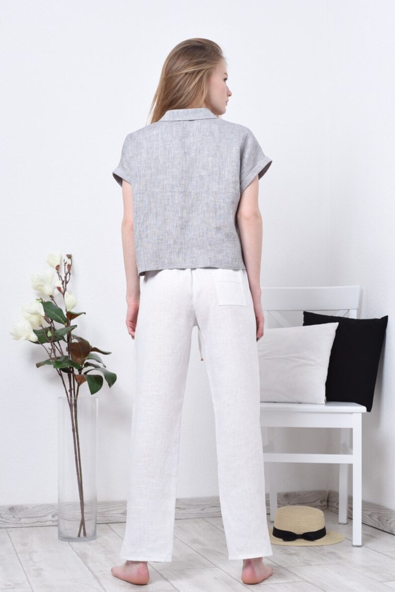 May include: A woman wearing a gray linen shirt with short sleeves and white linen pants. The pants have a drawstring waist and a pocket on the back.