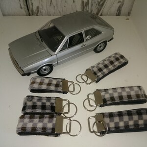 Puede incluir: Seis llaveros con tela a cuadros en blanco y negro y anillos de llaves plateados. Los llaveros están dispuestos en dos filas de tres. Un coche de juguete plateado está en el fondo.