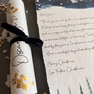 May include: A rolled up letter with gold glitter and a handwritten message that says "Hello Oscar. Thank you so much for the mince pies, beer and Rudolph was so happy that you had left him a such an easy one to find because of the reindeer. Reindeer smelt it from miles away! I've heard kind boy this year so I've delivered a newt. Have a wonderful day celebrating with Merry Christmas. Love Father Christmas x". The letter is on a white background with a blue watercolour background with stars and a silhouette of Santa's sleigh.