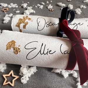 May include: Two white paper Christmas crackers with gold foil accents and black calligraphy. The crackers are tied together with a dark blue ribbon and a burgundy velvet ribbon. The text on the crackers reads "Oscar (age 2)" and "Ellie (age 3)".