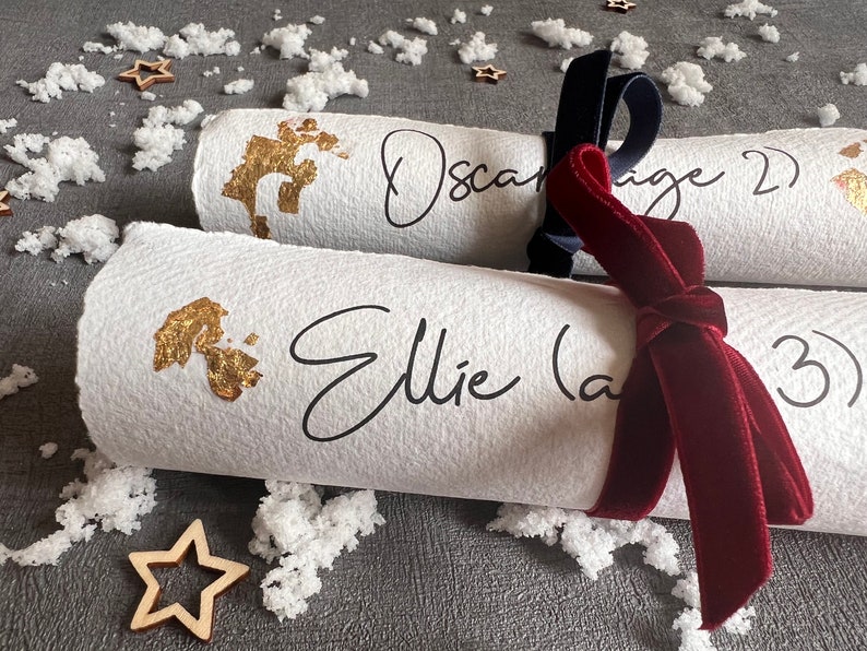 May include: Two white paper Christmas crackers with gold foil accents and black calligraphy. The crackers are tied together with a dark blue ribbon and a burgundy velvet ribbon. The text on the crackers reads "Oscar (age 2)" and "Ellie (age 3)".